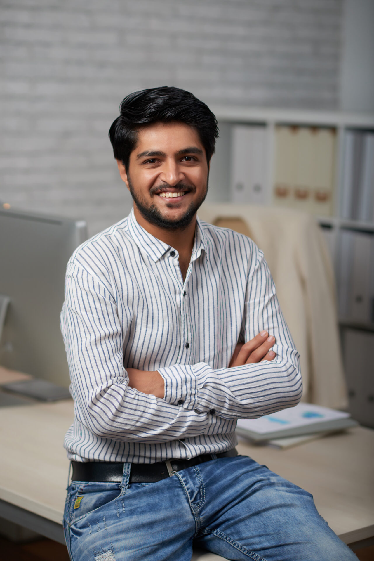 Confident young Indian businessman sitting on edge of his table and looking at camera